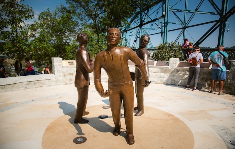 bronze sculpture of three people holding hands in a circle installed next to a pedestrian bridge