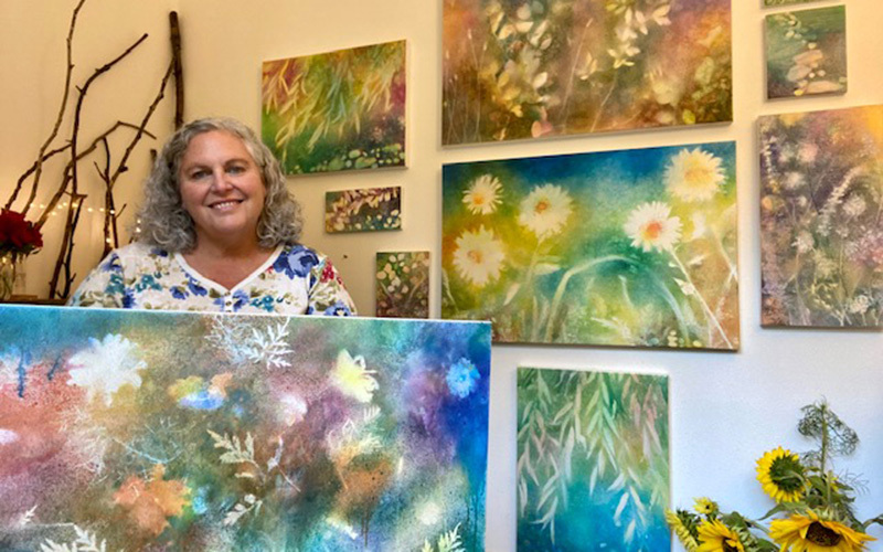 An artist holding up her painting against a backdrop of paintings