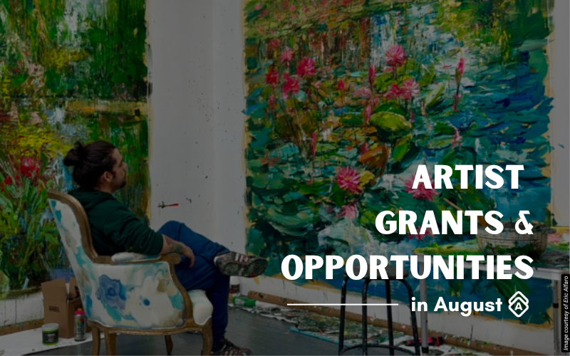 Artist Eric Alfaro sits in his studio observing large-scale landscape paintings displayed on white walls. He wears a dark green sweatshirt and is seated in a paint-splattered chair. The vibrant canvases feature lush natural scenes with thick, expressive brushwork depicting foliage, flowers in pinks and greens, and water elements in blues and teals. The impressionistic style captures botanical subjects with energetic, layered paint application. Art supplies and materials are scattered on the studio floor. The bright, spacious environment allows for contemplation and creation of these immersive nature-inspired works. Text overlay reads "ARTIST GRANTS & OPPORTUNITIES in August" in white letters on the right side of the image.