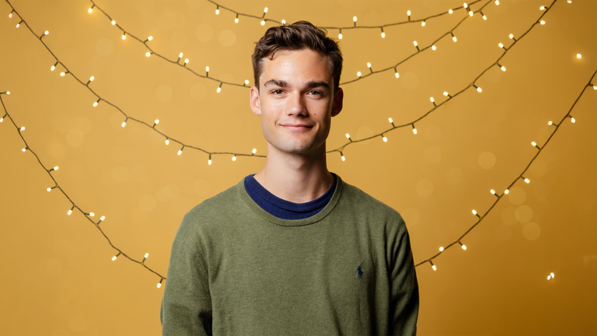 A person in a green sweater stands smiling in front of a yellow background with string lights.
