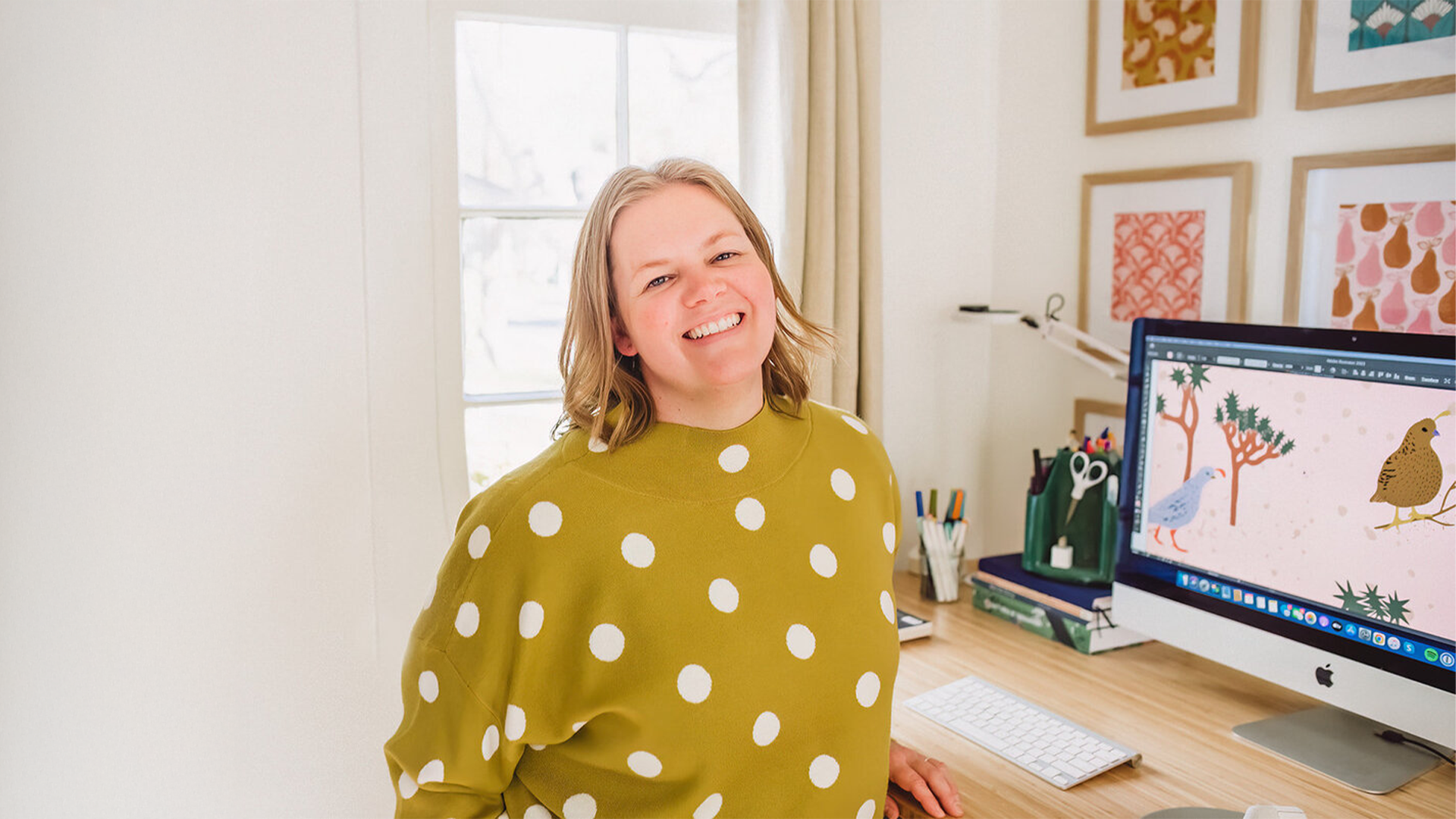Smiling person in a green polka dot sweater sits at a desk with a computer displaying colorful artwork, surrounded by framed pictures.
